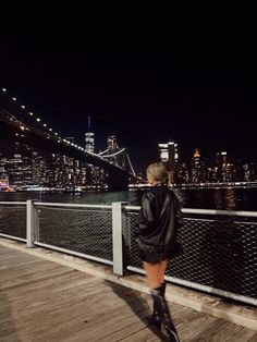 a woman standing on the side of a bridge looking at the water and city lights