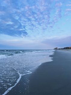 a beach with waves coming in to shore and houses on the other side, under a cloudy blue sky