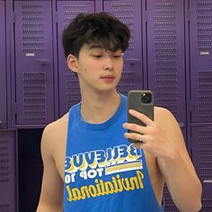 a young man in a blue tank top is looking at his cell phone while standing in front of lockers