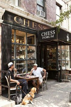 two men sitting at a table with their dogs in front of a chess forum store