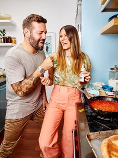 a man and woman standing in a kitchen with food on the stove, smiling at each other