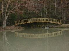 a wooden bridge over a body of water with trees in the background and one person sitting on a bench under it