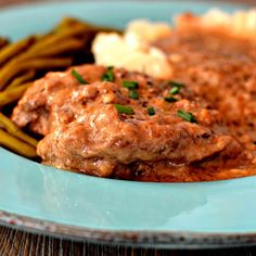 a blue plate topped with meat covered in gravy next to green beans and mashed potatoes