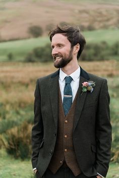 a man wearing a suit and tie standing in front of a green field with hills