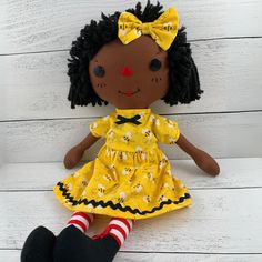 a doll with black hair wearing a yellow dress and striped socks sitting on a white wooden surface