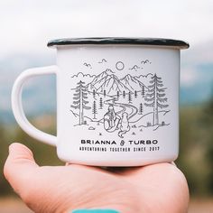 a hand holding a white coffee mug with a drawing on the front and side of it