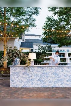 a woman standing behind a blue and white counter in front of a house with lights strung over it