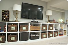 a tv mounted on top of a white shelf filled with boxes and baskets next to lamps