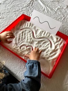 a young child is playing with sand in a red tray on top of a bed