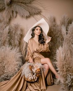 a woman in a long dress and hat sitting on a chair with her legs crossed