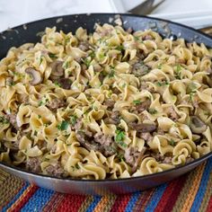 a skillet filled with pasta and meat on top of a colorful place mat next to silverware