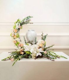 an arrangement of flowers and greenery on a table in front of a white vase