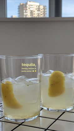 two glasses filled with liquid sitting on top of a tiled counter next to a window