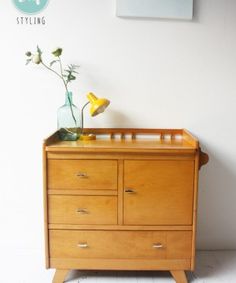 a wooden dresser with flowers in a glass vase on it's top, against a white wall