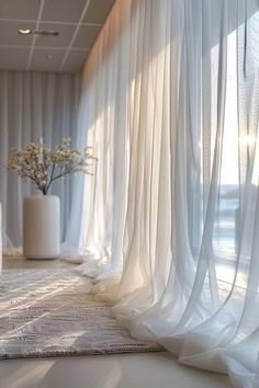 a white vase with flowers in it sitting on the floor next to a curtained wall