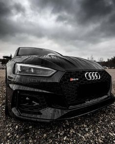 the front end of a black sports car on gravel with dark clouds in the background