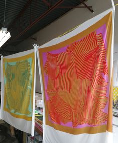 three colorful paintings hanging from the ceiling in an art studio, one with orange and pink colors