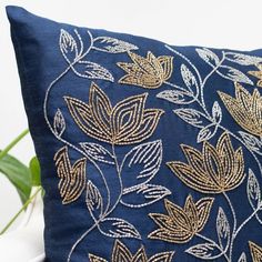 a blue pillow with gold leaf embroidered on the front and back, sitting next to a potted plant