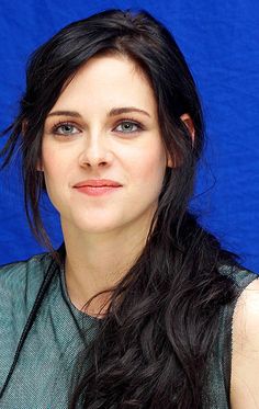 a woman with long black hair wearing a green top and posing for a photo in front of a blue backdrop