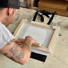 a man sitting at a table working on a piece of wood with an unfinished frame