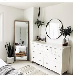 a bedroom with white furniture and plants on the dresser next to a large round mirror