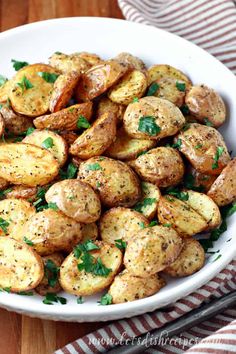 a white bowl filled with roasted potatoes on top of a wooden table next to a red and white striped napkin