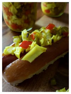 a hot dog covered in toppings sitting on top of a wooden table next to a jar of pickles