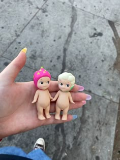 someone holding two small toy dolls in their hand on the sidewalk, one is wearing a pink hat