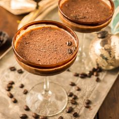 two glasses filled with chocolate drink sitting on top of a table next to coffee beans