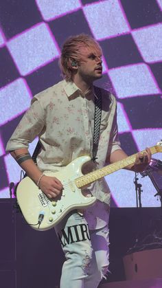 a man with blonde hair playing an electric guitar on stage at a music festival in front of a checkered background