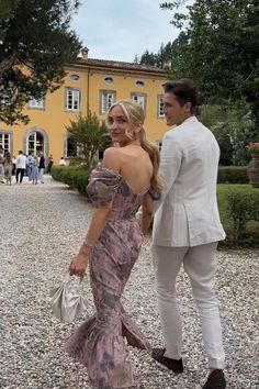 a man in a white suit and woman in a pink dress are standing on gravel