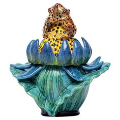 a frog sitting on top of a blue flower with green leaves and yellow spots in its mouth