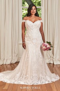 a woman standing in front of a window wearing a wedding dress with off the shoulder sleeves