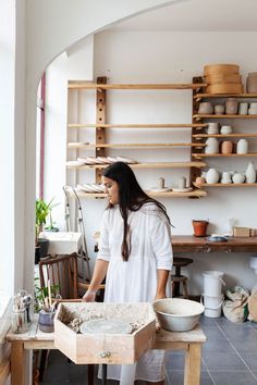 a woman standing in front of a wooden table filled with bowls and pottery on shelves