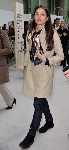 a woman in a trench coat and scarf walks through an exhibit area with other people