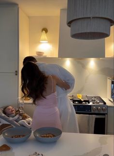 a woman in a pink dress holding a baby while standing next to a stove top oven