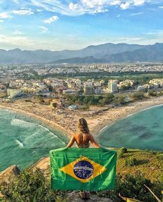 a woman sitting on top of a hill next to the ocean holding a brazil flag