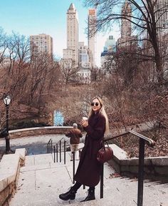 a woman is standing on the steps drinking coffee and looking at her cell phone in central park