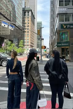 three women standing on the sidewalk in front of a crosswalk with cars and tall buildings behind them