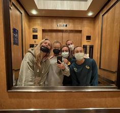 four women wearing face masks taking a selfie in front of a mirror with their cell phones