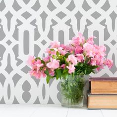 pink flowers are in a vase next to two books on a white table with a gray wallpaper behind it