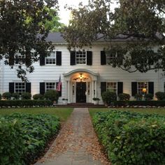 a large white house with black shutters on the front and side windows, surrounded by hedges