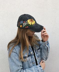 a woman wearing a denim jacket and hat with embroidered flowers on the side, leaning against a white wall