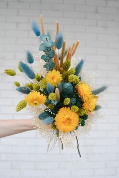 a bouquet of yellow and blue flowers is held by someone's hand in front of a brick wall