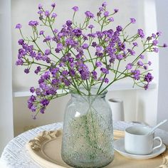 purple flowers are in a glass vase on a plate with a cup and saucer