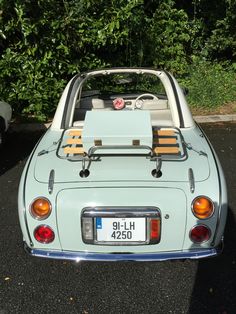 an old blue car parked in front of some bushes and trees with its hood up