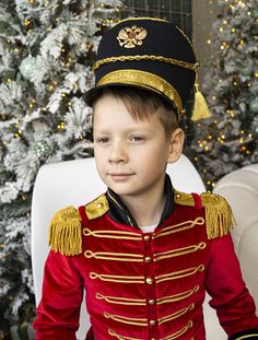 a young boy dressed in a red and gold uniform sitting next to a christmas tree