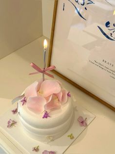 a white cake with pink flowers and a lit candle on top sits in front of a framed photograph