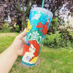 someone holding up a starbucks cup with ariel the mermaid on it in front of a tree