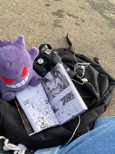 a purple stuffed animal sitting on top of a bag next to a book and camera